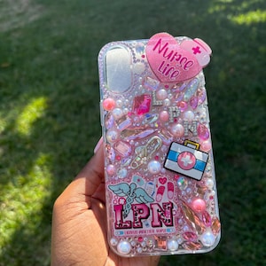 May include: A clear phone case decorated with pink and clear rhinestones, pearls, and charms. The case features a heart-shaped accent with the words "Nurse Life" and a medical symbol. Other charms include a medical bag and the letters "LPN".