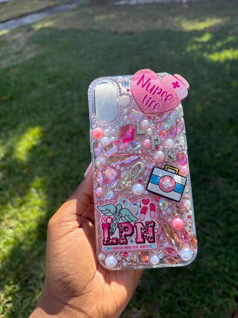 May include: A clear phone case decorated with pink and clear rhinestones, pearls, and charms. The case features a heart-shaped accent with the words "Nurse Life" and a medical symbol. Other charms include a medical bag and the letters "LPN".