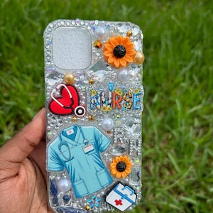 May include: Clear phone case decorated with rhinestones, pearls, and charms. The design includes a red heart with a stethoscope, a blue medical scrub illustration, orange sunflowers, and the word "NURSE".