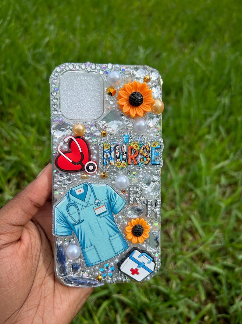 May include: Clear phone case decorated with rhinestones, pearls, and charms. The design includes a red heart with a stethoscope, a blue medical scrub illustration, orange sunflowers, and the word "NURSE".