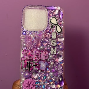 May include: A clear phone case bedazzled with purple and silver rhinestones, pearls, and charms. The case features a butterfly, a stethoscope, and the words "SCRUB LIFE" and "BSN". A purple flower and a small shirt charm are also visible.