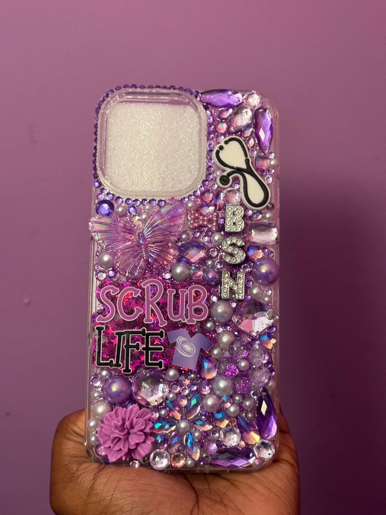 May include: A clear phone case bedazzled with purple and silver rhinestones, pearls, and charms. The case features a butterfly, a stethoscope, and the words "SCRUB LIFE" and "BSN". A purple flower and a small shirt charm are also visible.