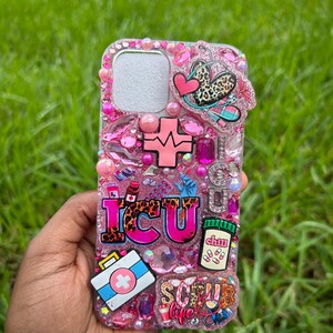 May include: Pink bedazzled phone case with the letters "ICU" in leopard print, a cross, a first aid kit, and a pill bottle. The case is decorated with pink gems, hearts, and the words "Scrub Life".