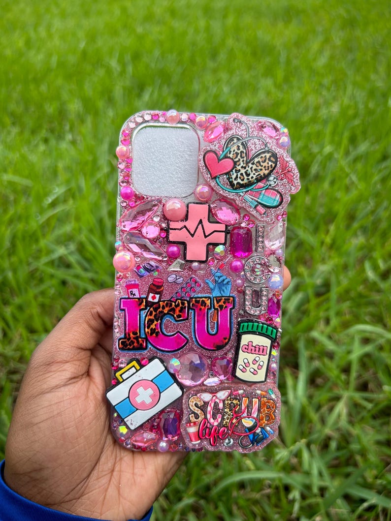 May include: Pink bedazzled phone case with the letters "ICU" in leopard print, a cross, a first aid kit, and a pill bottle. The case is decorated with pink gems, hearts, and the words "Scrub Life".