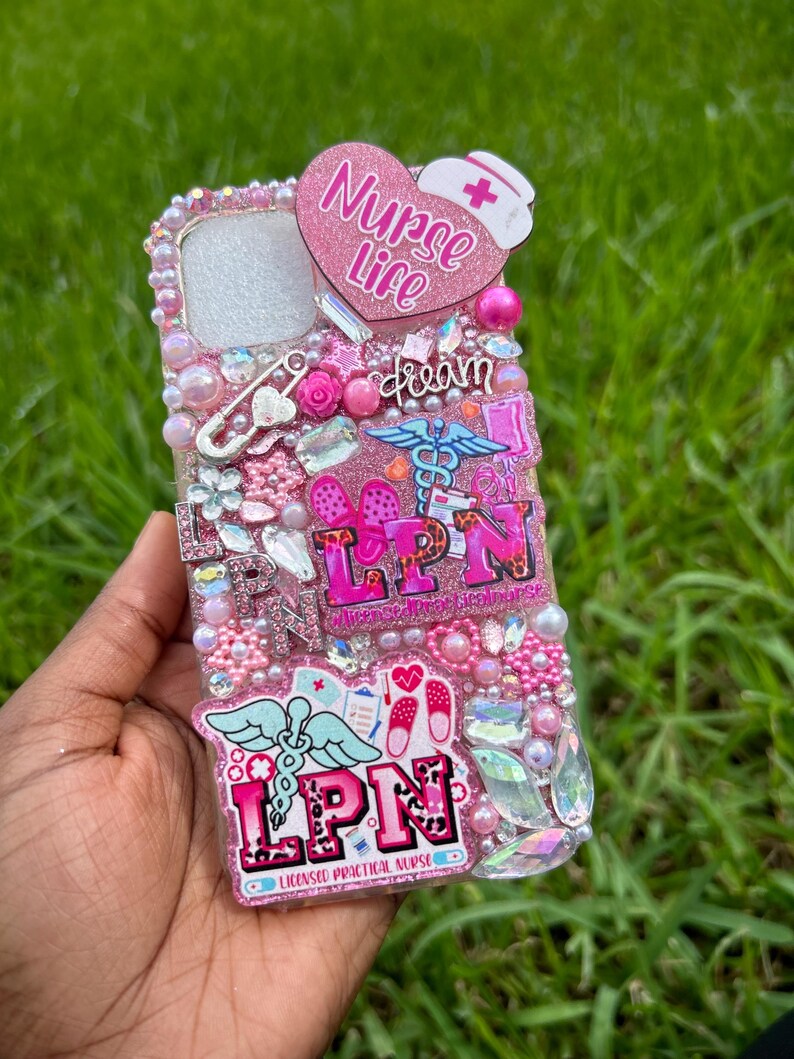 May include: A pink and white bedazzled phone case with the words "Nurse Life" in a heart shape. The case is decorated with pink pearls, rhinestones, and the letters "LPN". Additional embellishments include a nurse's hat, medical symbols, and the word "dream".