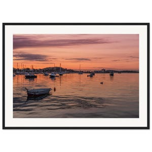May include: A framed landscape photograph of a harbor at sunset. The sky is filled with soft pink and orange clouds reflecting on the calm water. Several small boats and sailboats are anchored in the distance, with one small white boat in the foreground.