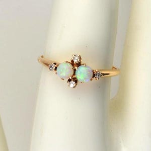 May include: A gold ring featuring two oval opals and small accent diamonds. The ring is set on a gold band and is displayed on a white surface. The opals have a milky white color.