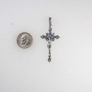 May include: A silver-toned crucifix pendant with a detailed depiction of Jesus Christ. The pendant is approximately 2 inches in length and features intricate detailing. A U.S. dime is included for size comparison. The pendant has a loop for a chain.