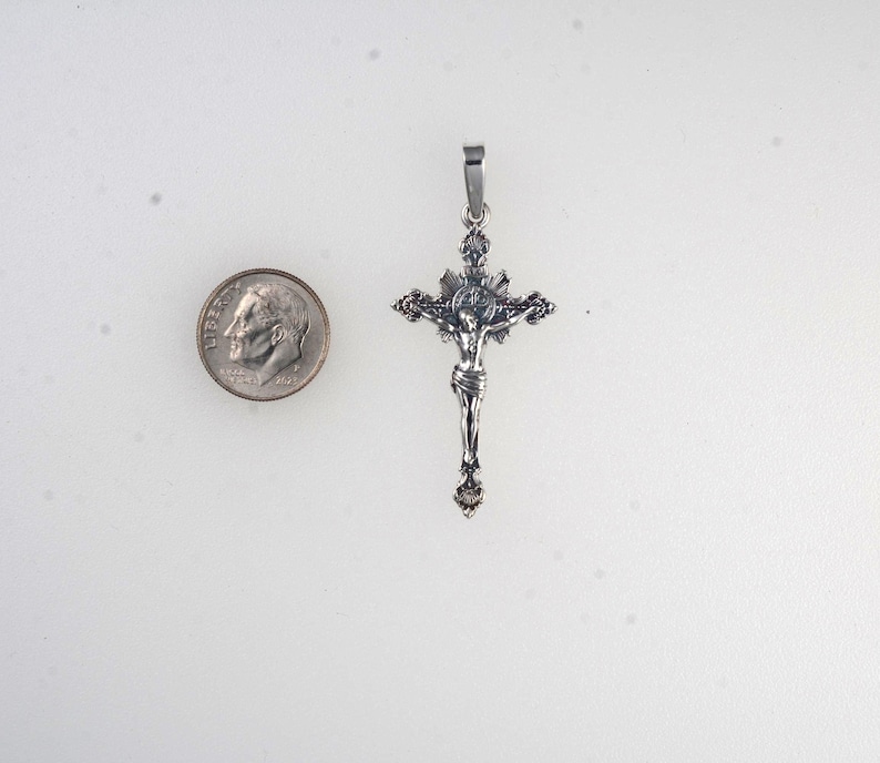 May include: A silver-toned crucifix pendant with a detailed depiction of Jesus Christ. The pendant is approximately 2 inches in length and features intricate detailing. A U.S. dime is included for size comparison. The pendant has a loop for a chain.