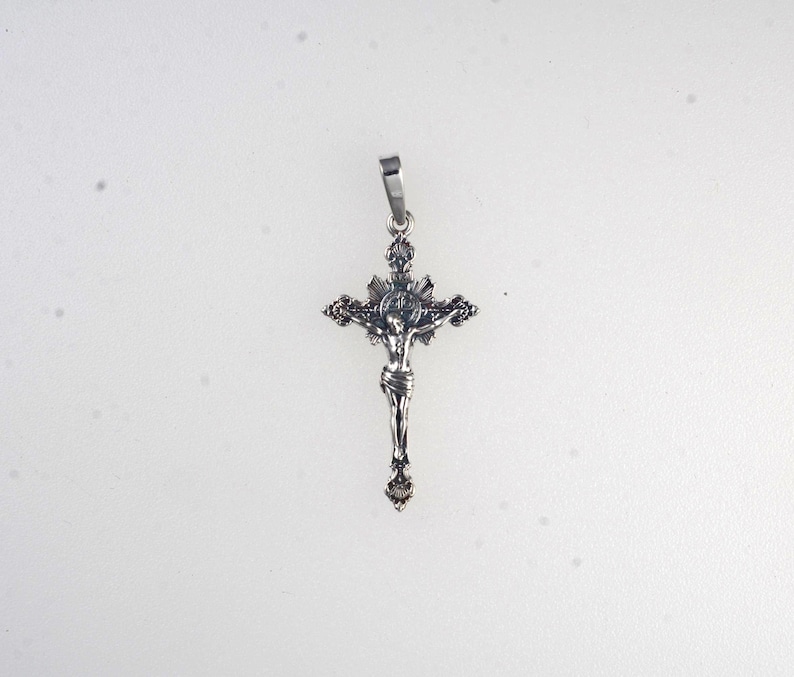 May include: A silver-colored crucifix pendant. The cross features a detailed depiction of Jesus Christ. The pendant has a loop at the top for a chain. The design includes intricate detailing on the cross arms and the figure of Christ.