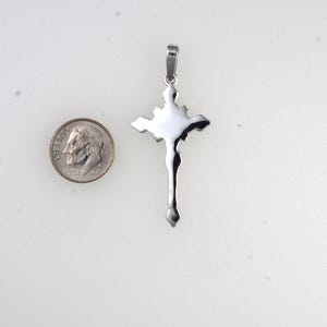 May include: A silver-colored pendant in the shape of a cross, with a detailed depiction of a figure. A U.S. dime is next to the pendant for size comparison. The cross has a loop at the top for a chain.