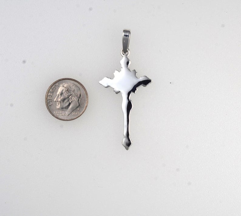 May include: A silver-colored pendant in the shape of a cross, with a detailed depiction of a figure. A U.S. dime is next to the pendant for size comparison. The cross has a loop at the top for a chain.