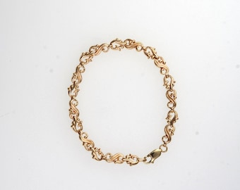 May include: A gold-colored bracelet with an ornate, repeating scroll design. The bracelet is made of linked segments, each featuring a stylized scroll pattern. A clasp secures the bracelet. The jewelry is displayed against a white background.