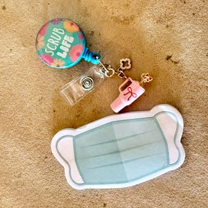 May include: A teal and floral "SCRUB LIFE" badge reel with a clear plastic ID holder, a pink charm, and a light blue face mask sticker on a tan surface. The badge reel has a retractable cord.