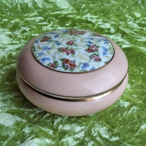 Könnte beinhalten: Eine runde, hellrosa Schmuckschatulle mit einem Blumendesign auf dem Deckel. Der Deckel hat einen weißen Hintergrund mit rosa, blauen und orangefarbenen Blumen. Ein goldenes Band umrandet den Deckel und den Boden der Schatulle.