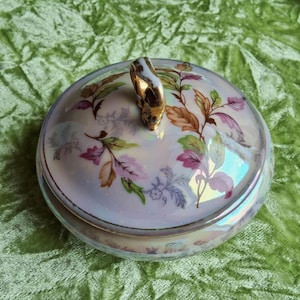 Könnte beinhalten: Eine runde, schillernde Schmuckdose aus Keramik mit Deckel. Der Deckel hat einen goldenen Griff und ist mit einem Blumenmuster in Rosa-, Grün- und Brauntönen verziert. Die Dose steht auf einer grünen Oberfläche.