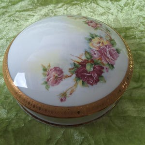 Könnte beinhalten: Eine weiße Porzellan-Schmuckdose mit goldfarbenem Rand und einem Blumendesign mit rosa und roten Rosen. Der Deckel ist gewölbt und die Dose ist rund.