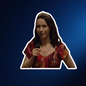 Op de afbeelding: Een persoon met donker haar draagt een tiara en een jurk met korte mouwen in een rood en oranje patroon, terwijl deze een zwarte microfoon vasthoudt. De figuur is wit omlijnd tegen een donkerblauwe achtergrond tijdens een spreekmoment.