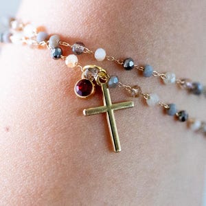 May include: A gold cross pendant hangs from a delicate beaded bracelet. The bracelet features small, multi-coloured beads and a small, round, red gemstone. The jewelry is designed to be worn on the wrist.