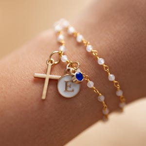 May include: Gold-toned bracelet featuring a cross charm and a circular charm with the letter "E". The bracelet has a double strand design with small white beads and a blue accent. The bracelet is worn on a wrist.