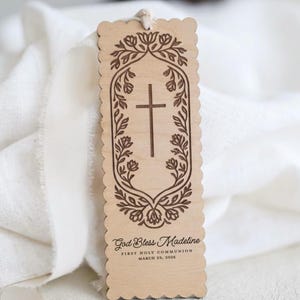 May include: A rectangular light wood bookmark with scalloped edges and a white string at the top. It features an engraved cross surrounded by a floral border, with the text God Bless Madeline, FIRST HOLY COMMUNION, and MARCH 29, 2026 engraved below.
