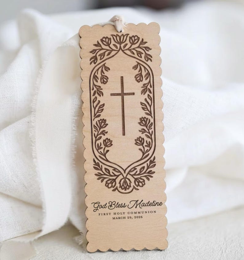 May include: A rectangular light wood bookmark with scalloped edges and a white string at the top. It features an engraved cross surrounded by a floral border, with the text God Bless Madeline, FIRST HOLY COMMUNION, and MARCH 29, 2026 engraved below.