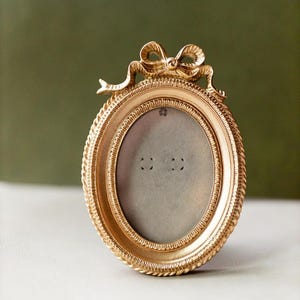 Puede incluir: Un marco de fotos ovalado dorado con un lazo decorativo en la parte superior. El marco tiene un borde en forma de cuerda y un interior de color neutro. El marco está sobre una superficie blanca con un fondo verde.