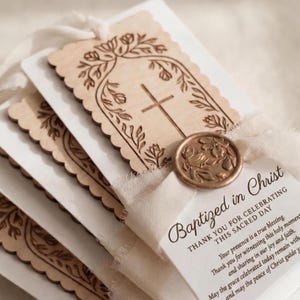May include: A stack of rectangular baptism favor tags featuring light wood cutouts with scalloped edges, engraved with a cross and floral arch. Each tag is tied with cream-colored ribbon and a bronze-toned wax seal with a floral design.