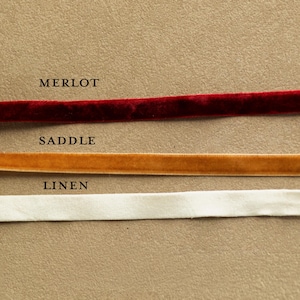 May include: Close-up of three velvet ribbons in Merlot, Saddle, and Linen colors. The Merlot ribbon is a deep red, the Saddle ribbon is a warm gold, and the Linen ribbon is a soft white. The names of the colors are printed above each ribbon.