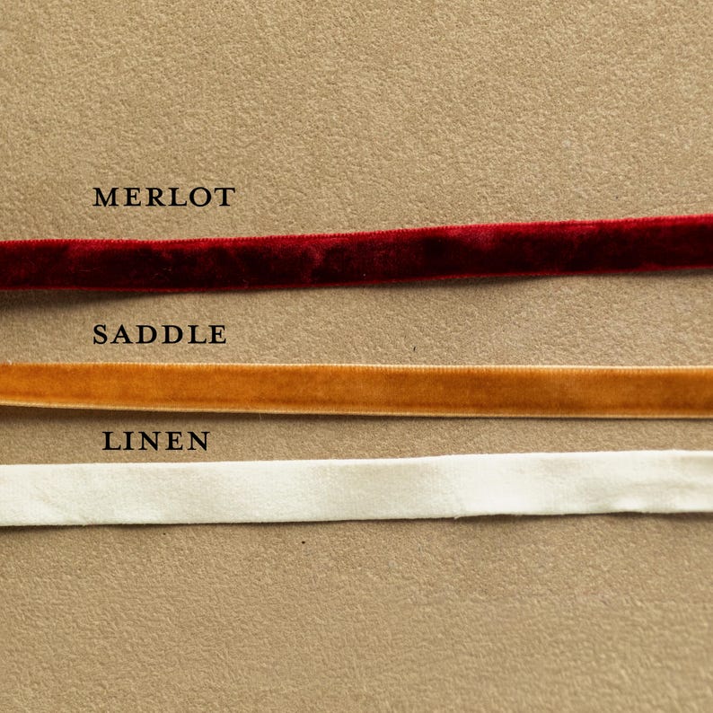 May include: Close-up of three velvet ribbons in Merlot, Saddle, and Linen colors. The Merlot ribbon is a deep red, the Saddle ribbon is a warm gold, and the Linen ribbon is a soft white. The names of the colors are printed above each ribbon.