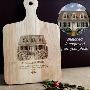 May include: A light-coloured wooden chopping board with a handle, engraved with a house illustration and text. The text reads "THE MILLER HOME" and a Bible verse. A circular image shows a house. The text "sketched & engraved from your photo" is also present.