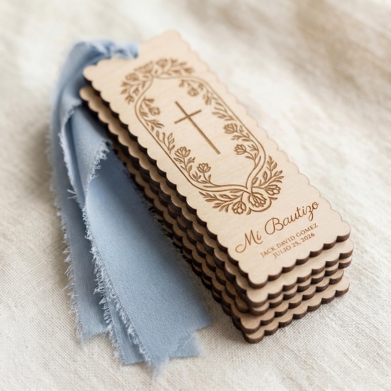 May include: A stack of rectangular wooden bookmarks with scalloped edges, each featuring an engraved cross surrounded by a floral border. The text reads Mi Bautizo, Jack David Gomez, and July 25, 2026. A dusty blue frayed ribbon is attached to the top.