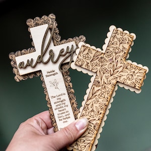 May include: Two wooden crosses with floral and leaf patterns. One cross has the name "Audrey" in a script font. The other cross has a scalloped edge. One cross has the text "The Lord is my strength and my shield; my heart trusts in him, and he helps me."