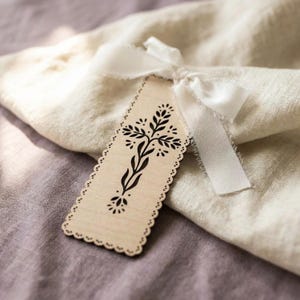 May include: A rectangular light-colored wooden tag with scalloped edges and a black printed floral cross design. The tag is tied with a white fabric ribbon featuring frayed edges, resting atop a folded piece of light beige textured fabric.