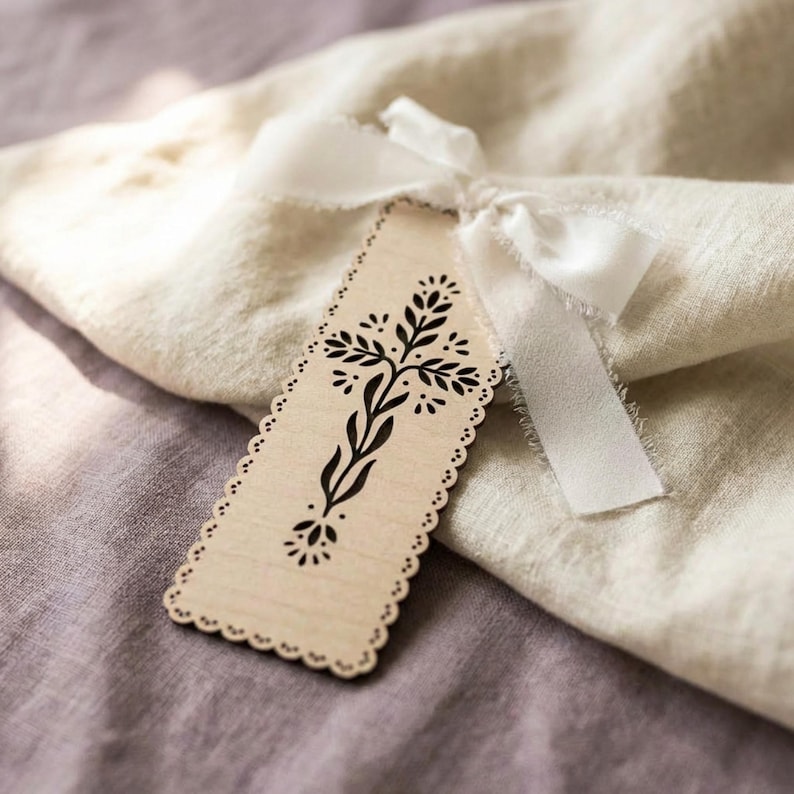 May include: A rectangular light-colored wooden tag with scalloped edges and a black printed floral cross design. The tag is tied with a white fabric ribbon featuring frayed edges, resting atop a folded piece of light beige textured fabric.