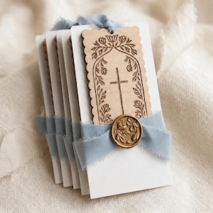 May include: A stack of rectangular white cardstock tags, each featuring a scalloped-edge wooden topper engraved with a cross and floral arch. A dusty blue frayed ribbon secures the wood to the card, finished with a circular gold wax seal.