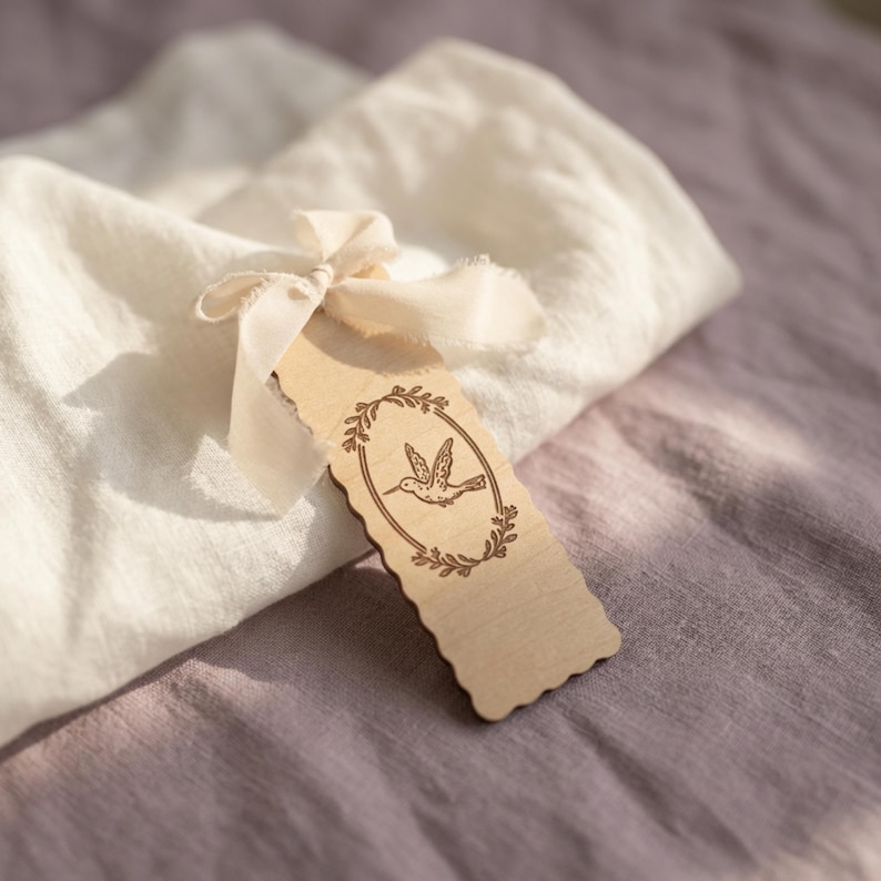 May include: A rectangular light-colored wooden bookmark with scalloped edges, featuring a laser-engraved illustration of a hummingbird centered within an oval laurel wreath. A cream-colored fabric ribbon is tied through a hole at the top.