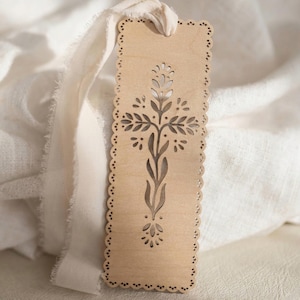May include: A rectangular light brown wooden bookmark with a scalloped edge and a laser-cut cross design featuring stylized leaves and petals. A long, frayed, off-white fabric ribbon is threaded through a hole at the top of the wooden bookmark.
