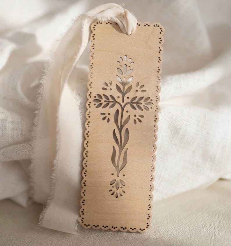 May include: A rectangular light brown wooden bookmark with a scalloped edge and a laser-cut cross design featuring stylized leaves and petals. A long, frayed, off-white fabric ribbon is threaded through a hole at the top of the wooden bookmark.