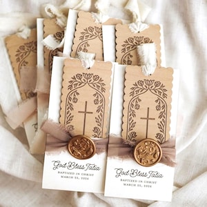 May include: Rectangular white cardstock tags featuring a scalloped-edge light brown wooden overlay with a laser-engraved cross and floral arch. Each tag is tied with a white ribbon, a taupe fabric strip, and a gold wax seal. Text reads God Bless Talia.