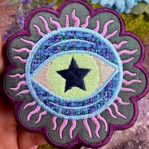 May include: Embroidered patch featuring an eye design with a star in the center. The eye is surrounded by a blue and purple design, with pink sun-like rays. The patch has a gray background and a purple scalloped border.