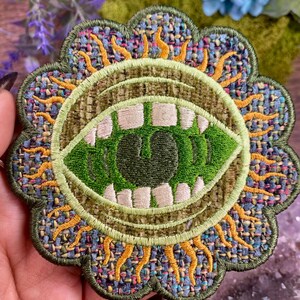 May include: Embroidered patch featuring a green, open-mouthed design with white teeth and a green interior. The patch has a floral-shaped border with a woven texture in shades of blue, green, and yellow. The design is reminiscent of a stylized eye or mouth.