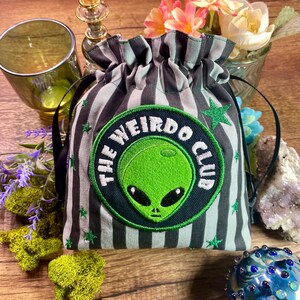 Puede incluir: Una bolsa con cordón con un patrón de rayas negras y grises y un parche circular con una cara de alienígena verde y las palabras "THE WEIRDO CLUB". La bolsa tiene un patrón de estrellas verdes y cordones negros. La bolsa está sobre una superficie de madera.