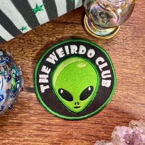 Könnte beinhalten: Ein runder Aufnäher mit dem Text "THE WEIRDO CLUB" in weißer Schrift um einen grünen Alienkopf. Der Aufnäher hat einen schwarzen Rand und liegt auf einer Holzoberfläche.