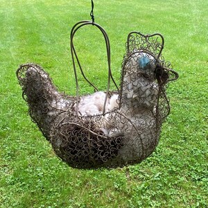 May include: A handcrafted wire chicken-shaped basket with a handle, filled with fluffy white material. The chicken has a detailed wire mesh body and a blue eye. The basket is suspended against a backdrop of green grass.