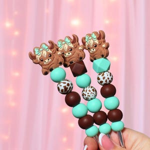 Puede incluir: Tres brochetas decorativas con diseño de vaca de las Tierras Altas. Cada brocheta presenta un dije de vaca marrón con un lazo azul, alternando con cuentas marrones, moteadas blancas y turquesas. Fondo rosa.