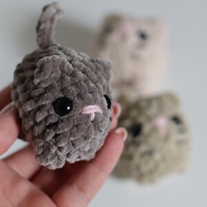 May include: A handmade gray plush cat with black eyes and a pink embroidered nose and mouth. The cat has a small tail and is held in a hand. Two other plush cats are in the background, one tan and one gray.