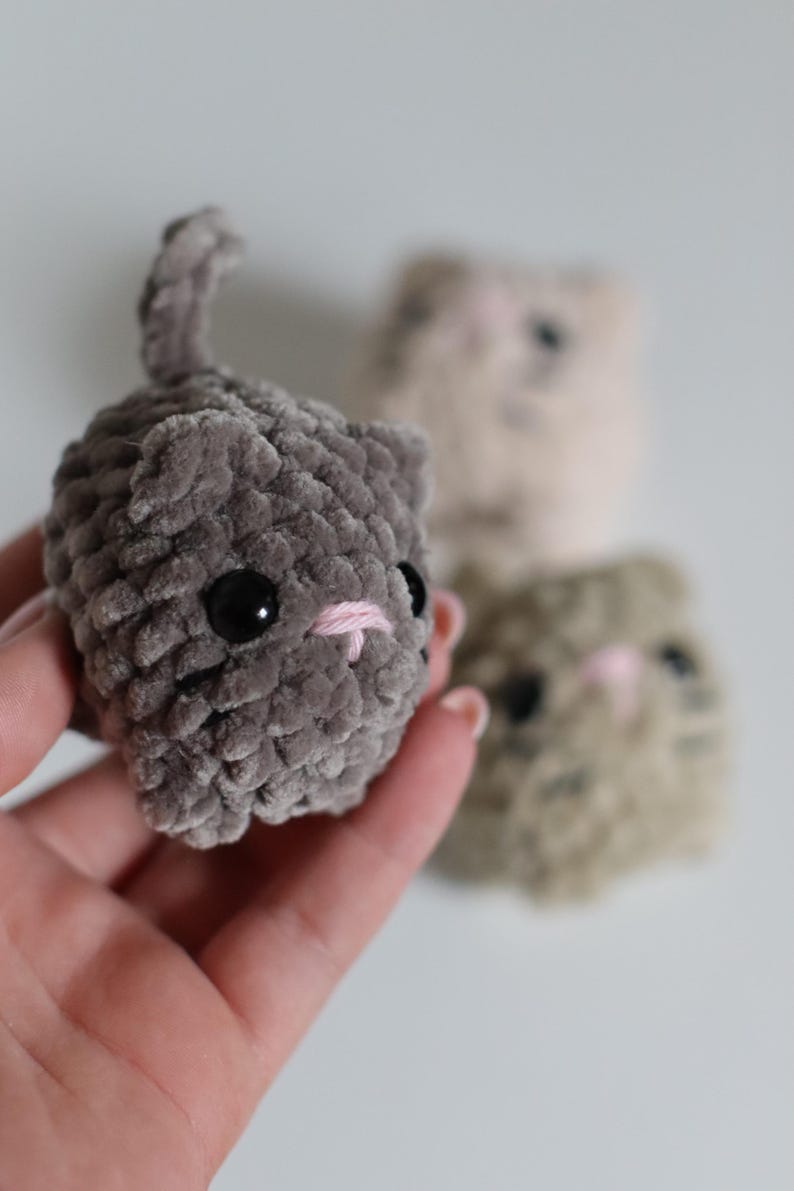May include: A handmade gray plush cat with black eyes and a pink embroidered nose and mouth. The cat has a small tail and is held in a hand. Two other plush cats are in the background, one tan and one gray.