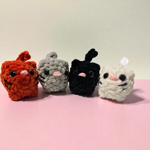 May include: Four crocheted cat toys in orange, gray, black, and white. Each cat has large black eyes, a pink nose, and black whiskers. The orange cat has a small, upright ear, while the black cat has a curved ear. The toys are on a pink surface.