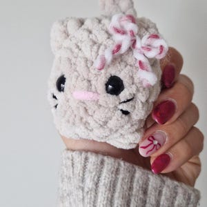 May include: A handmade, light gray crocheted cat-shaped item with black eyes, whiskers, and a pink nose. It has a small bow with red and white stripes. The item is held in a hand.